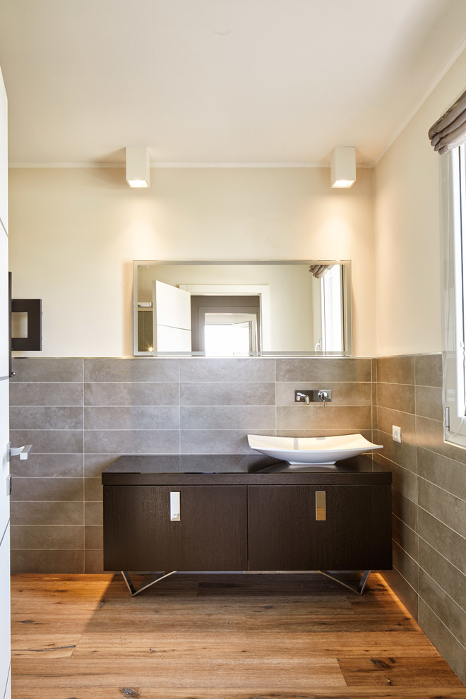 Idéer för att renovera ett funkis brun brunt badrum med dusch, med släta luckor, skåp i mörkt trä, grå kakel, porslinskakel, mellanmörkt trägolv, ett fristående handfat, bänkskiva i glas och flerfärgat golv