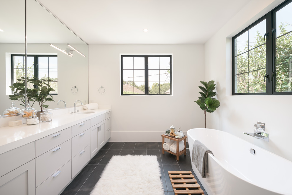 Пример оригинального дизайна: ванная комната в стиле неоклассика (современная классика) с фасадами в стиле шейкер, серыми фасадами, отдельно стоящей ванной, белыми стенами, врезной раковиной, черным полом и белой столешницей