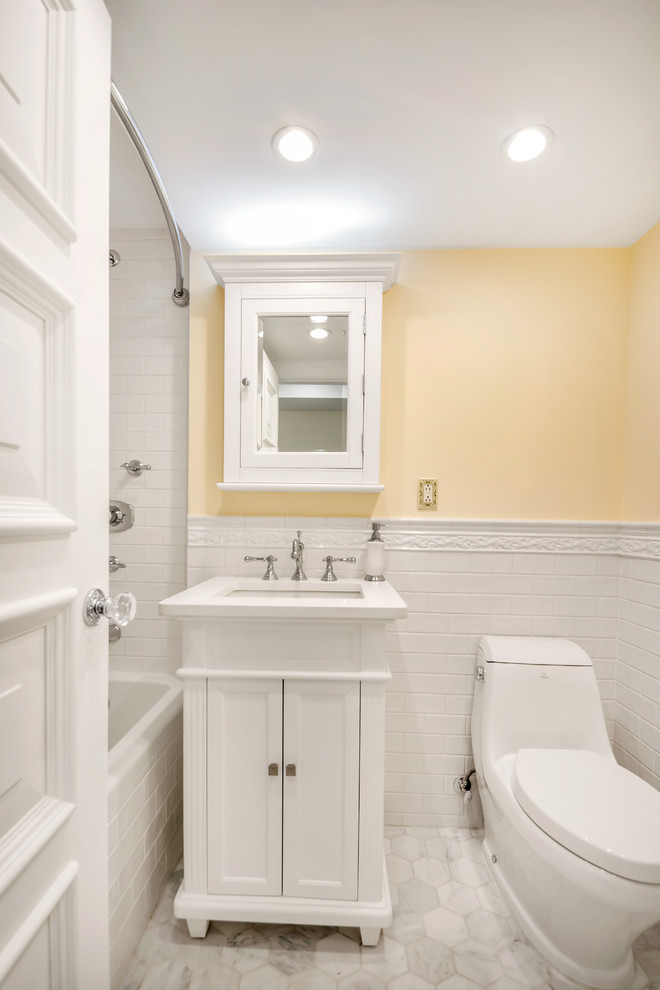 Bild på ett litet vintage badrum, med möbel-liknande, en toalettstol med hel cisternkåpa, vit kakel, stenkakel, vita skåp, marmorgolv, bänkskiva i akrylsten, ett badkar i en alkov, en dusch/badkar-kombination, gula väggar och ett integrerad handfat