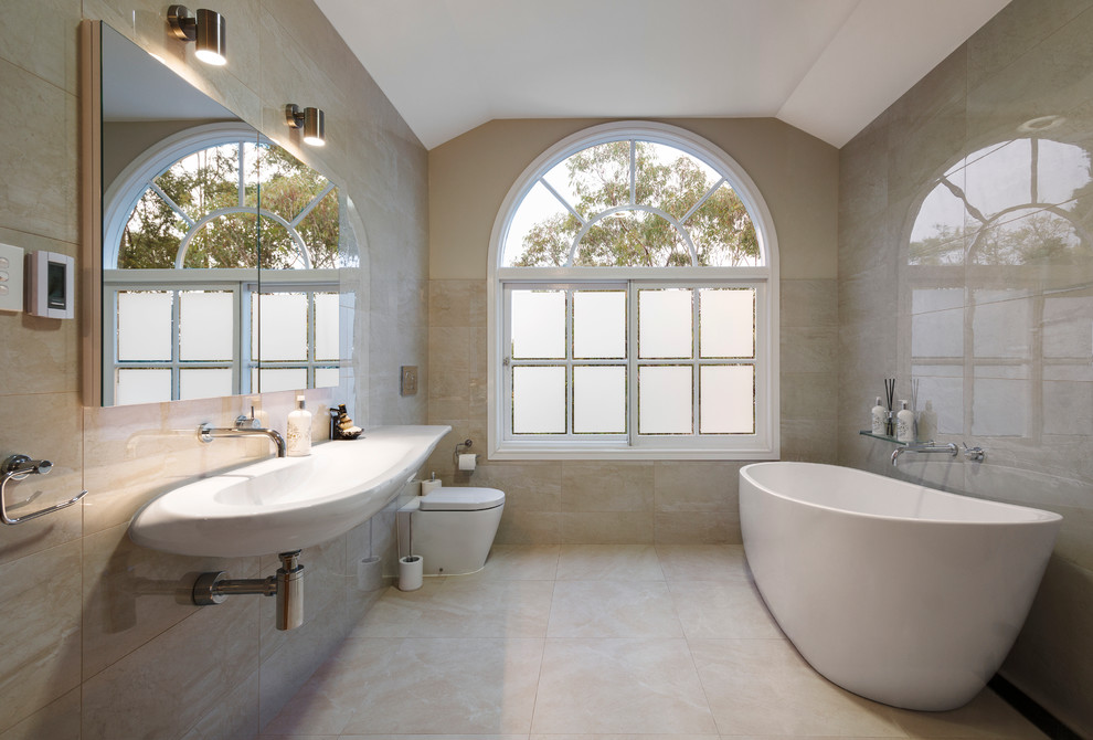 Klassisk inredning av ett stort en-suite badrum, med luckor med glaspanel, ett fristående badkar, en vägghängd toalettstol, beige kakel, beige väggar och ett väggmonterat handfat