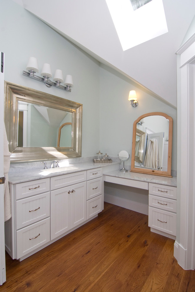 Cette image montre une grande salle de bain principale traditionnelle avec un placard à porte shaker, des portes de placard blanches, une baignoire en alcôve, un carrelage gris, un carrelage de pierre, un mur bleu, un sol en bois brun, un lavabo encastré et un plan de toilette en marbre.