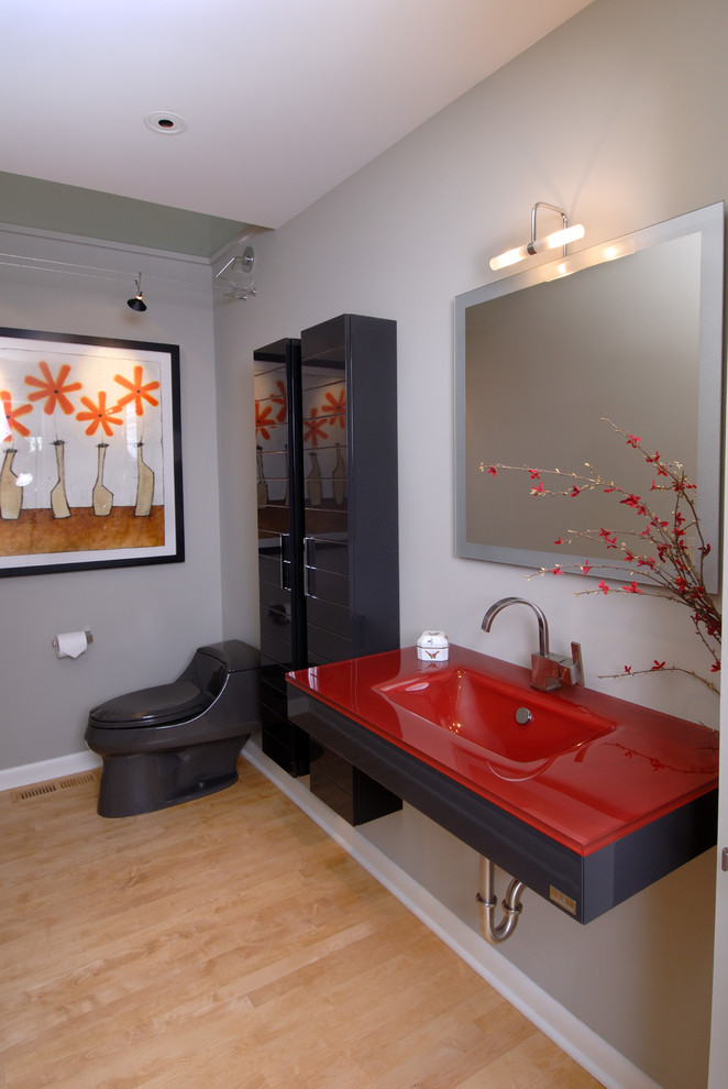 Ejemplo de cuarto de baño actual de tamaño medio con sanitario de una pieza, paredes grises, suelo de madera clara, lavabo integrado, encimera de vidrio, armarios con paneles lisos y puertas de armario negras