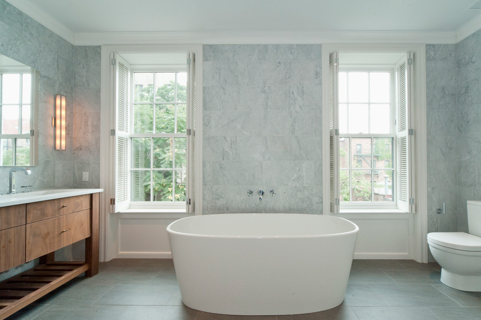 Cette photo montre une salle de bain principale chic en bois brun avec une baignoire indépendante, un carrelage blanc, un carrelage de pierre, WC à poser, un mur gris, un sol en carrelage de porcelaine, un lavabo encastré, un plan de toilette en quartz, un sol gris et un placard à porte plane.
