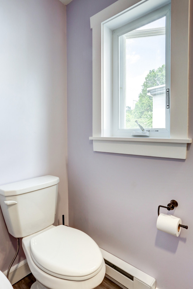 Cette image montre un WC et toilettes craftsman de taille moyenne avec des portes de placard beiges, un mur violet, un sol en vinyl, un lavabo encastré, un plan de toilette en marbre, un sol marron, un plan de toilette beige et meuble-lavabo sur pied.