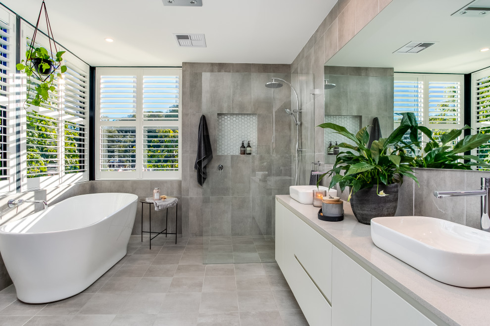 На фото: ванная комната в современном стиле с плоскими фасадами, белыми фасадами, отдельно стоящей ванной, серой плиткой, настольной раковиной, серым полом, серой столешницей и тумбой под две раковины