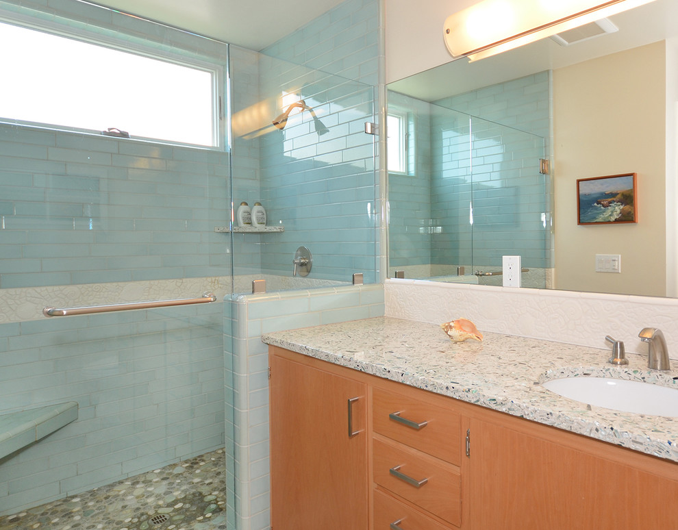 Bild på ett maritimt badrum med dusch, med ett undermonterad handfat, släta luckor, skåp i ljust trä, bänkskiva i återvunnet glas, en dusch i en alkov, blå kakel, porslinskakel, vita väggar och travertin golv