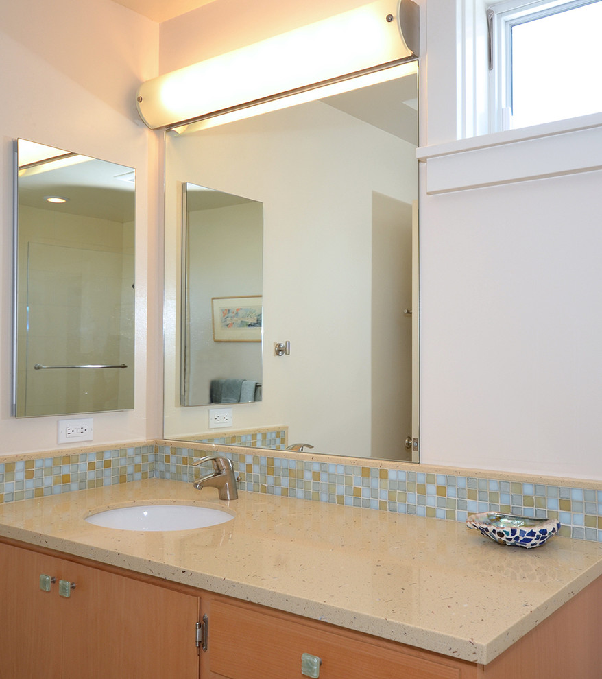 Cette photo montre une salle de bain bord de mer en bois clair avec un lavabo encastré, un placard à porte plane, un plan de toilette en verre recyclé, un carrelage bleu, un mur blanc et un sol en travertin.