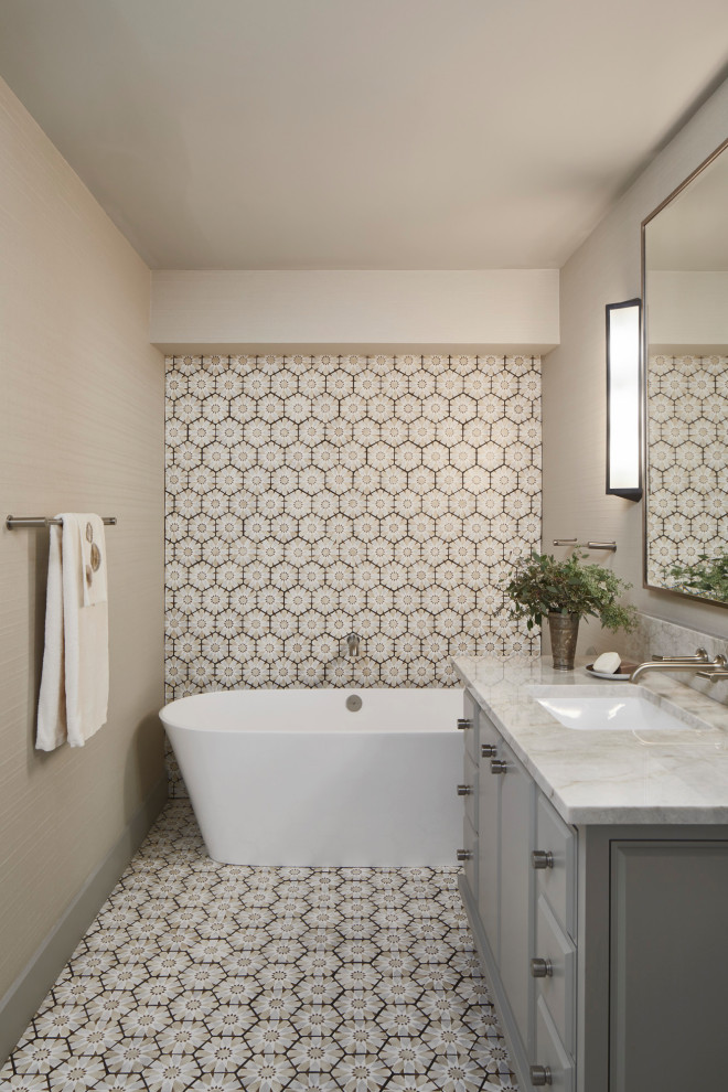 Cette image montre une salle de bain traditionnelle avec des portes de placard grises, une baignoire indépendante, un carrelage beige, un mur gris, un lavabo encastré, un sol multicolore et un plan de toilette gris.