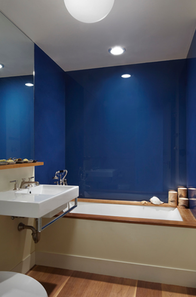 Idée de décoration pour une salle de bain principale design de taille moyenne avec un lavabo suspendu, une baignoire encastrée, un carrelage bleu, des plaques de verre, un sol en bois brun, un mur bleu, un placard sans porte et un sol marron.