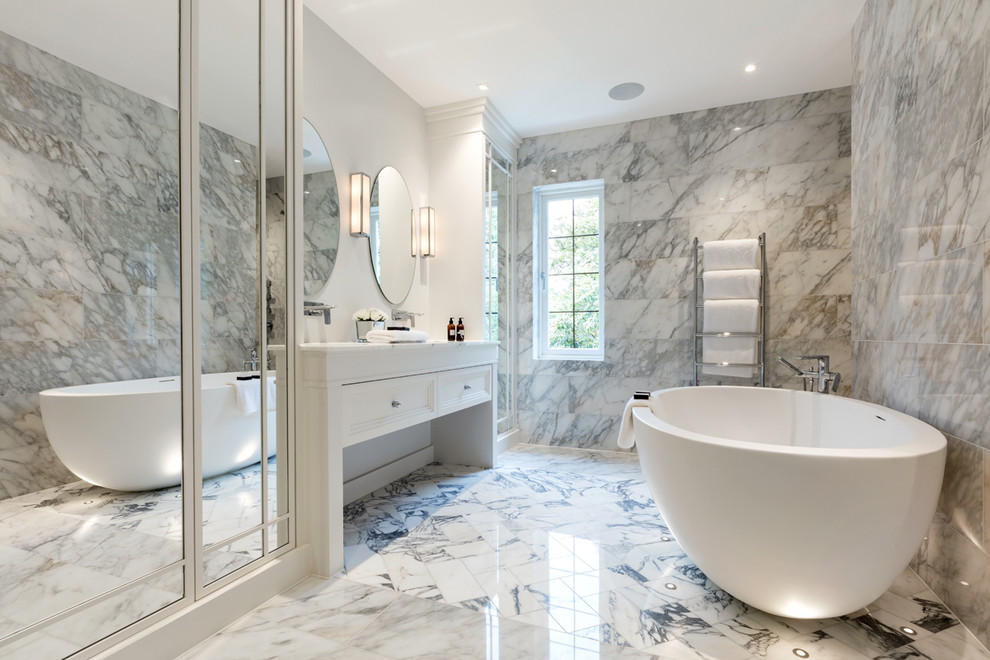 На фото: большая главная ванная комната в стиле неоклассика (современная классика) с белыми фасадами, отдельно стоящей ванной, серой плиткой, белой плиткой, белыми стенами, фасадами с утопленной филенкой и мраморным полом с
