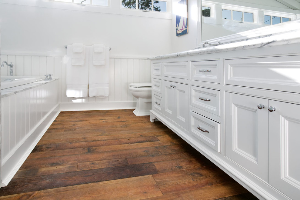 Example of a mid-sized beach style master medium tone wood floor drop-in bathtub design in Orange County with an undermount sink, recessed-panel cabinets, white cabinets, marble countertops, a two-piece toilet and white walls
