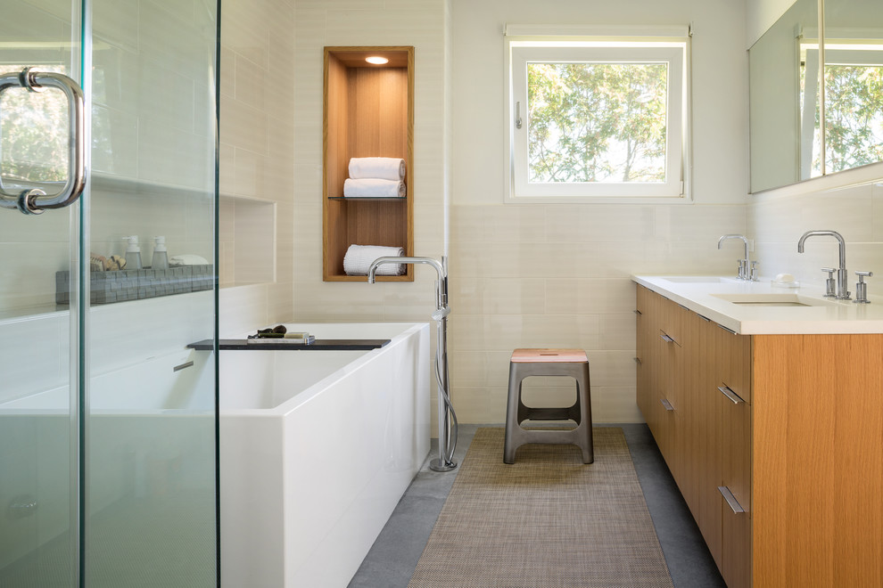 Modernes Badezimmer mit flächenbündigen Schrankfronten, hellbraunen Holzschränken, freistehender Badewanne, beigen Fliesen, weißer Wandfarbe, Betonboden, Unterbauwaschbecken, grauem Boden, Falttür-Duschabtrennung und weißer Waschtischplatte in Boston