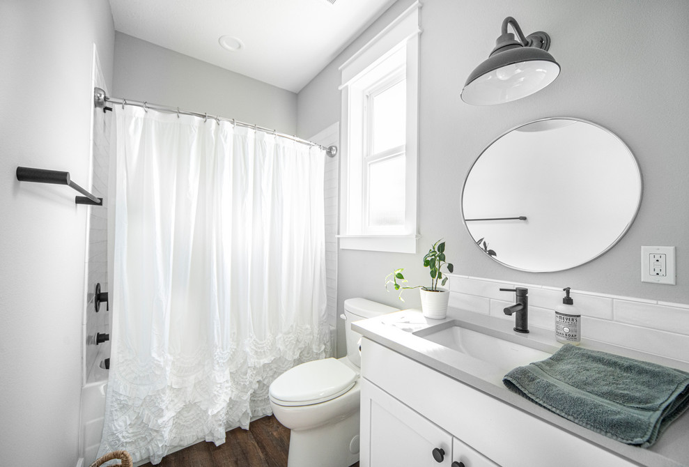 Klassisk inredning av ett mellanstort grå grått badrum med dusch, med skåp i shakerstil, vita skåp, ett badkar i en alkov, en dusch/badkar-kombination, en toalettstol med separat cisternkåpa, grå väggar, mellanmörkt trägolv, ett undermonterad handfat, bänkskiva i kvarts, brunt golv och dusch med duschdraperi