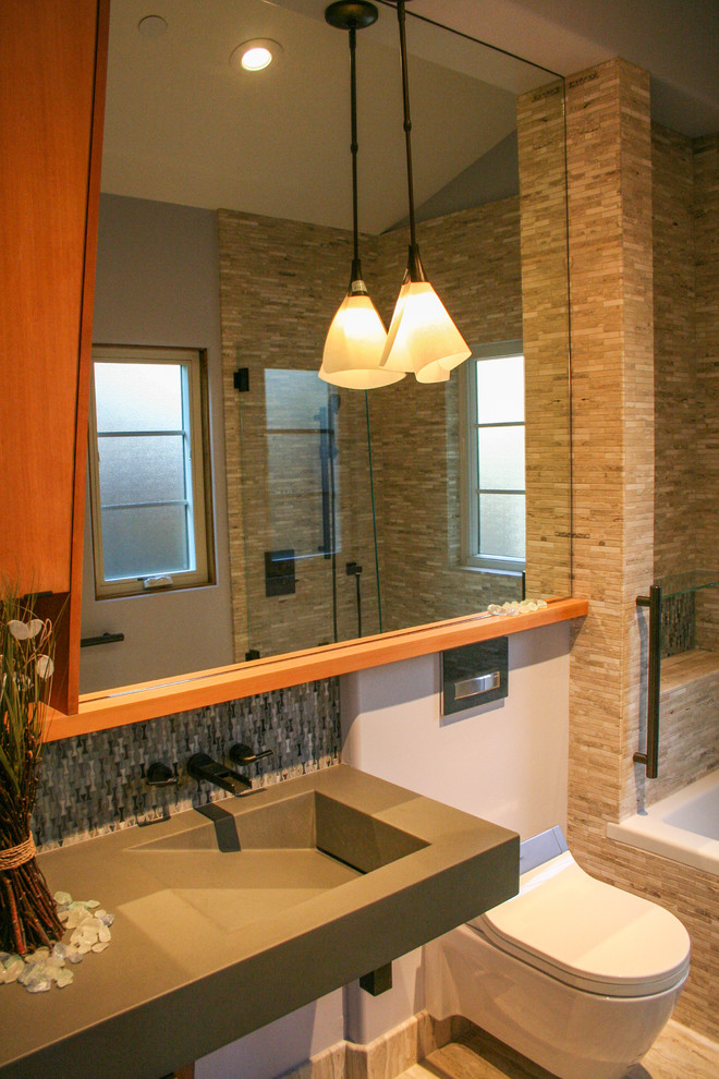 Modelo de cuarto de baño contemporáneo de tamaño medio con lavabo integrado, encimera de cemento, bañera encastrada, combinación de ducha y bañera, sanitario de pared, armarios con paneles lisos, puertas de armario de madera clara y baldosas y/o azulejos de piedra