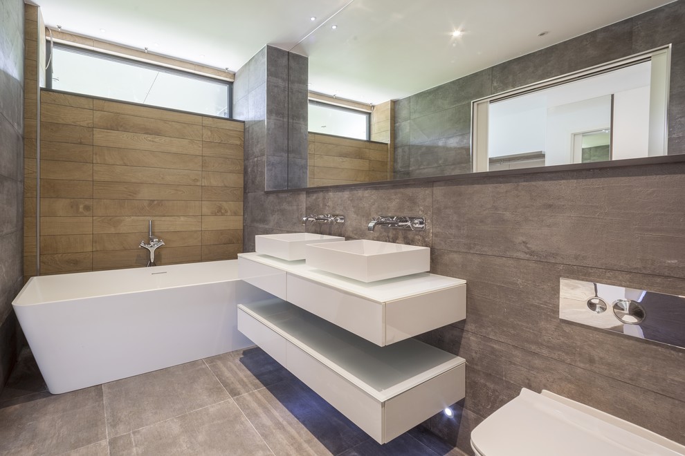 Photo of a contemporary bathroom in Dorset with flat-panel cabinets, white cabinets, a freestanding bath, a wall mounted toilet, grey tiles, a vessel sink, grey floors and white worktops.