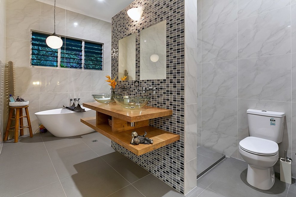 Пример оригинального дизайна: главная ванная комната среднего размера в современном стиле с открытыми фасадами, светлыми деревянными фасадами, отдельно стоящей ванной, раздельным унитазом, серой плиткой, разноцветной плиткой, серыми стенами, настольной раковиной, столешницей из дерева и серым полом