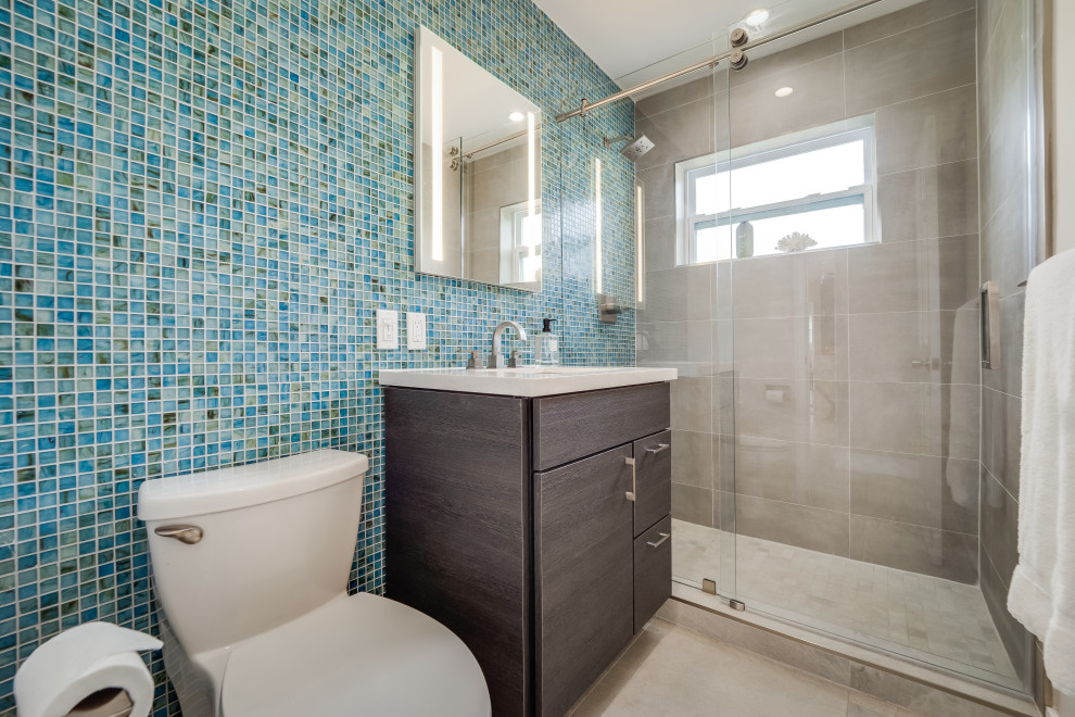 На фото: ванная комната среднего размера в стиле модернизм с плоскими фасадами, темными деревянными фасадами, душем в нише, унитазом-моноблоком, синей плиткой, плиткой из листового стекла, серыми стенами, полом из керамогранита, душевой кабиной, врезной раковиной, столешницей из искусственного кварца, серым полом, душем с раздвижными дверями, серой столешницей, тумбой под одну раковину и напольной тумбой