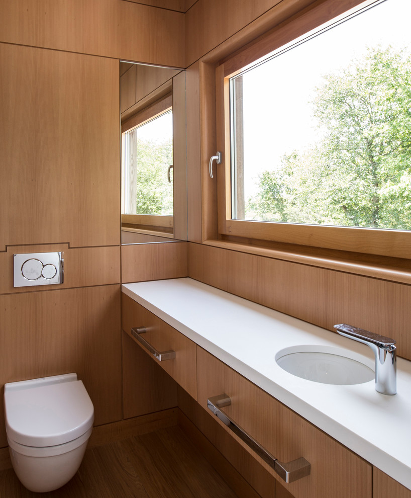 Imagen de cuarto de baño moderno pequeño con armarios con paneles lisos, puertas de armario de madera oscura, sanitario de pared, suelo de madera en tonos medios, lavabo bajoencimera y encimera de acrílico