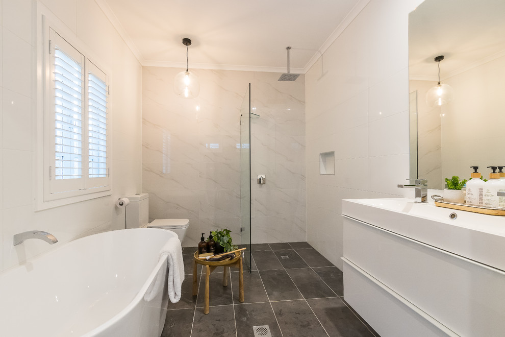 Inredning av ett klassiskt vit vitt badrum med dusch, med släta luckor, vita skåp, ett fristående badkar, våtrum, en toalettstol med separat cisternkåpa, vit kakel, ett integrerad handfat, grått golv och med dusch som är öppen