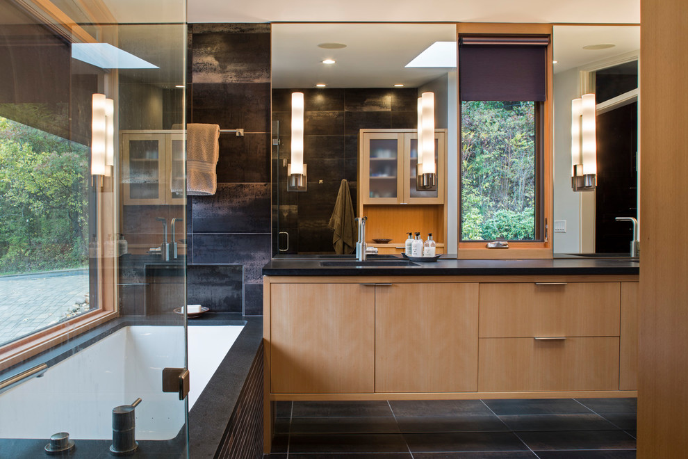 Cette photo montre une grande salle de bain principale tendance en bois clair avec une baignoire encastrée, un plan de toilette en granite, un placard à porte plane, un lavabo encastré, un carrelage noir, des carreaux de céramique, un mur beige, un sol en carrelage de céramique et un sol noir.