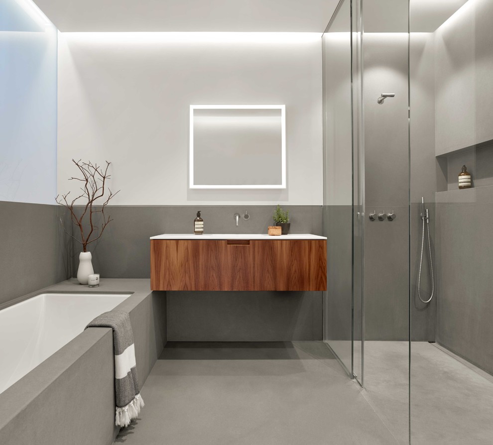 Modern inredning av ett mellanstort en-suite badrum, med möbel-liknande, skåp i mellenmörkt trä, ett platsbyggt badkar, en kantlös dusch, en vägghängd toalettstol, stenhäll, grå väggar, kalkstensgolv, ett integrerad handfat, bänkskiva i akrylsten, grått golv och dusch med skjutdörr