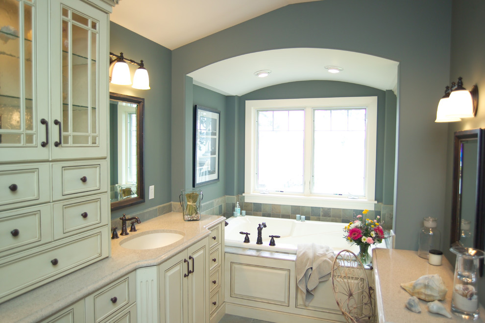 Photo of a large classic ensuite bathroom in Grand Rapids with recessed-panel cabinets, distressed cabinets, a built-in bath, a two-piece toilet, multi-coloured tiles, stone tiles, blue walls, a submerged sink and quartz worktops.