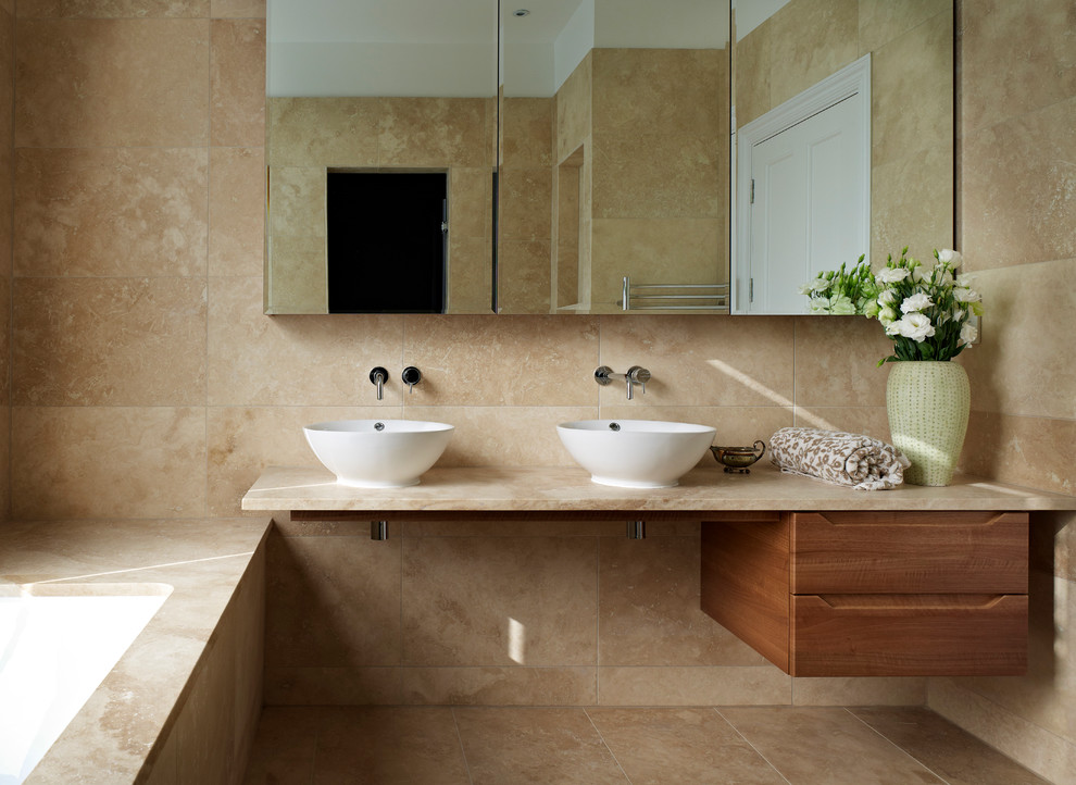 Источник вдохновения для домашнего уюта: ванная комната среднего размера в современном стиле с фасадами цвета дерева среднего тона, столешницей из известняка, настольной раковиной, полновстраиваемой ванной, бежевой плиткой и плоскими фасадами