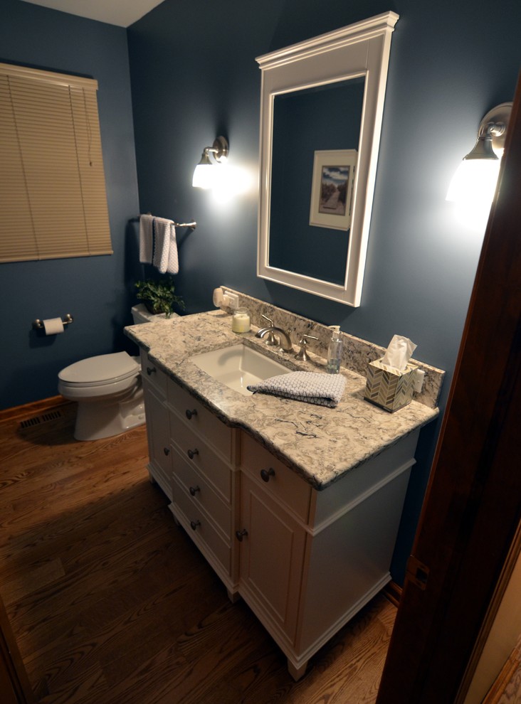 Cette image montre une petite salle d'eau traditionnelle avec un lavabo encastré, un placard avec porte à panneau encastré, des portes de placard blanches, un plan de toilette en quartz modifié, un mur bleu et un sol en bois brun.
