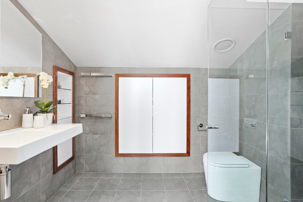 This is an example of a small contemporary ensuite bathroom in Sydney with grey tiles, ceramic tiles, ceramic flooring, a wall-mounted sink, grey floors, a hinged door, recessed-panel cabinets, white cabinets, a corner shower, a wall mounted toilet and glass worktops.