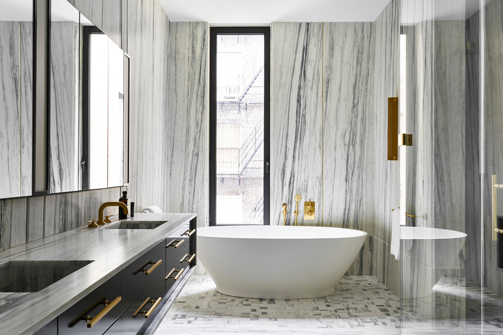 Cette image montre une salle de bain design avec un placard à porte plane, des portes de placard noires, une baignoire indépendante, un carrelage gris, un carrelage blanc, des dalles de pierre, un mur blanc, un sol en carrelage de terre cuite, un lavabo intégré, un sol blanc et un plan de toilette gris.
