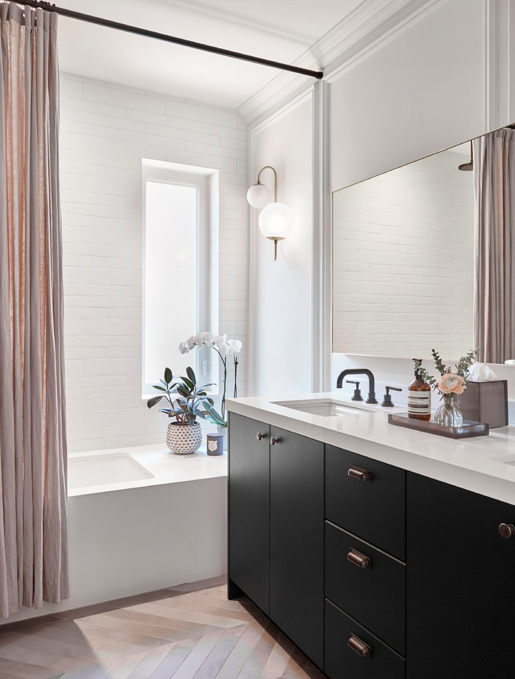 Cette image montre une salle de bain traditionnelle avec un placard à porte plane, des portes de placard noires, une baignoire encastrée, un carrelage blanc, un mur blanc, un lavabo encastré, un sol beige et un plan de toilette blanc.