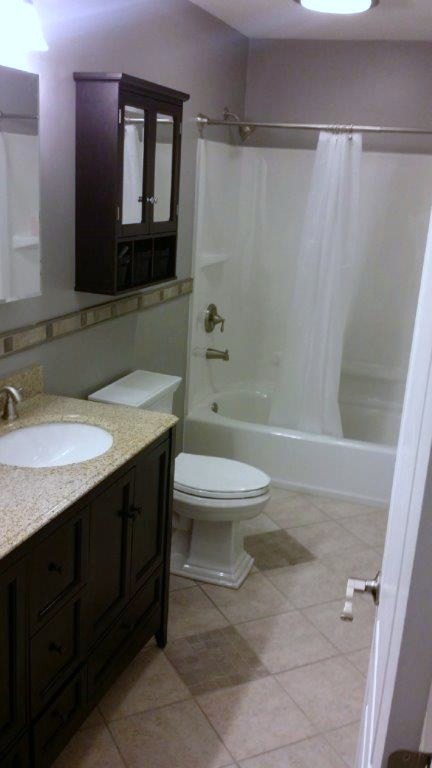 Klassisk inredning av ett litet badrum, med ett undermonterad handfat, skåp i shakerstil, skåp i mörkt trä, granitbänkskiva, ett badkar i en alkov, en dusch/badkar-kombination, en toalettstol med separat cisternkåpa, grå kakel, porslinskakel, grå väggar och klinkergolv i porslin