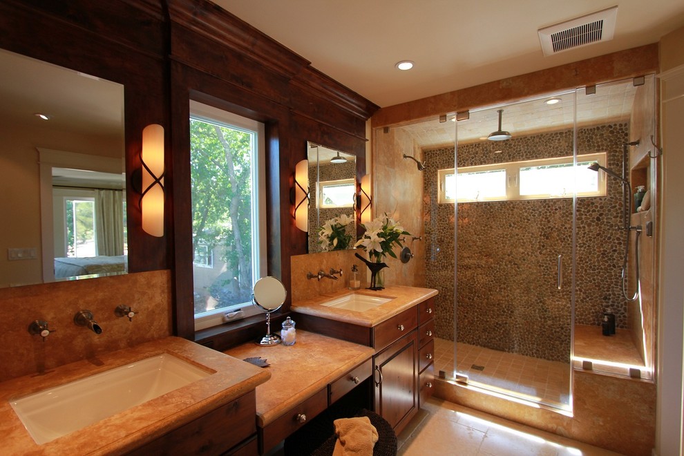 Idée de décoration pour une grande salle de bain craftsman en bois brun avec un lavabo encastré, un placard avec porte à panneau surélevé, un plan de toilette en calcaire, WC à poser, un carrelage beige, une plaque de galets et un sol en calcaire.