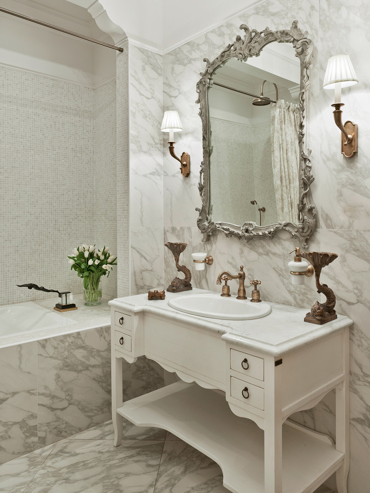 Klassisk inredning av ett en-suite badrum, med ett nedsänkt handfat, vita skåp, en dusch/badkar-kombination, vit kakel, ett badkar i en alkov, stenhäll, vita väggar, marmorgolv och släta luckor