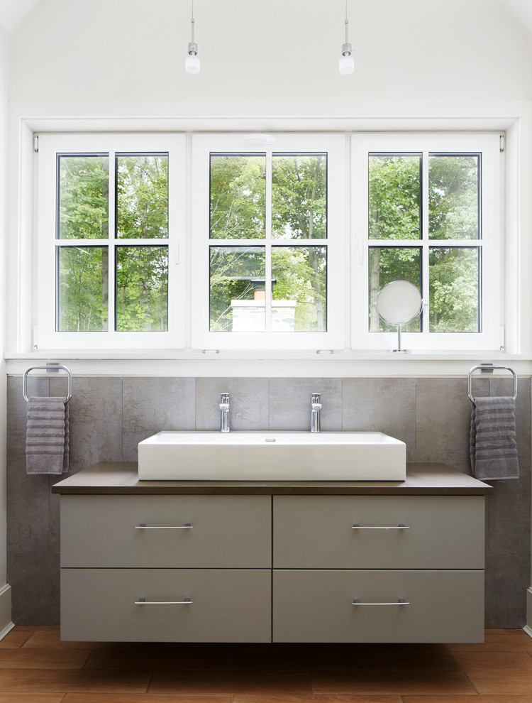 Imagen de cuarto de baño contemporáneo con armarios con paneles lisos, puertas de armario grises, baldosas y/o azulejos grises, paredes blancas, suelo de madera en tonos medios y lavabo de seno grande