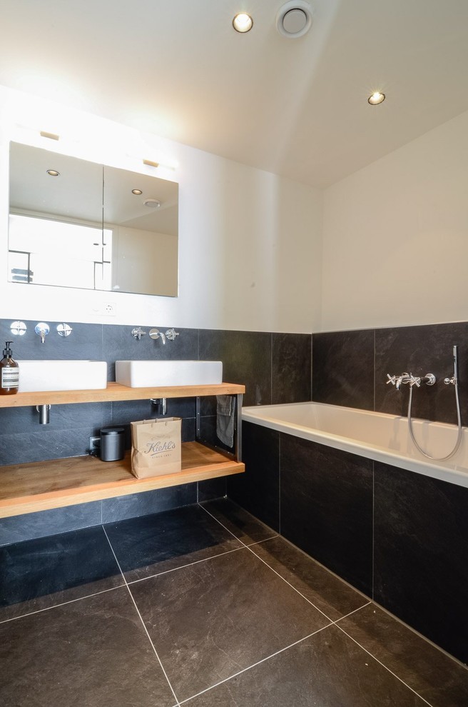 Inspiration for a medium sized contemporary ensuite bathroom in Other with a vessel sink, open cabinets, light wood cabinets, wooden worktops, a shower/bath combination, a wall mounted toilet, black tiles, white walls, ceramic flooring and a built-in bath.