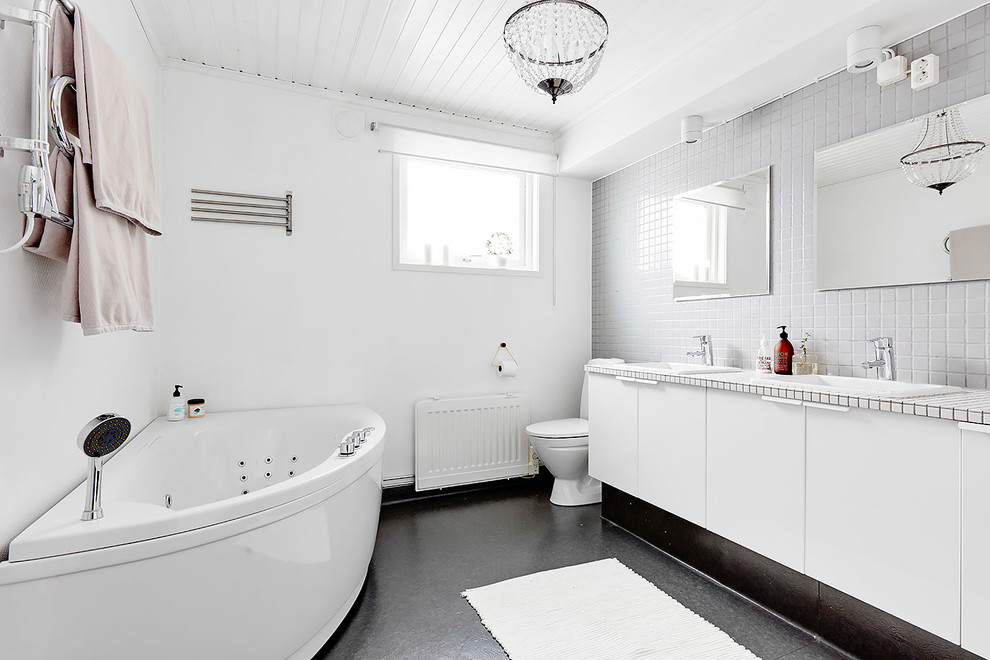 На фото: большая главная ванная комната в скандинавском стиле с белыми фасадами, угловой ванной, душем в нише, унитазом-моноблоком, белой плиткой, белыми стенами, накладной раковиной, столешницей из плитки и плоскими фасадами с