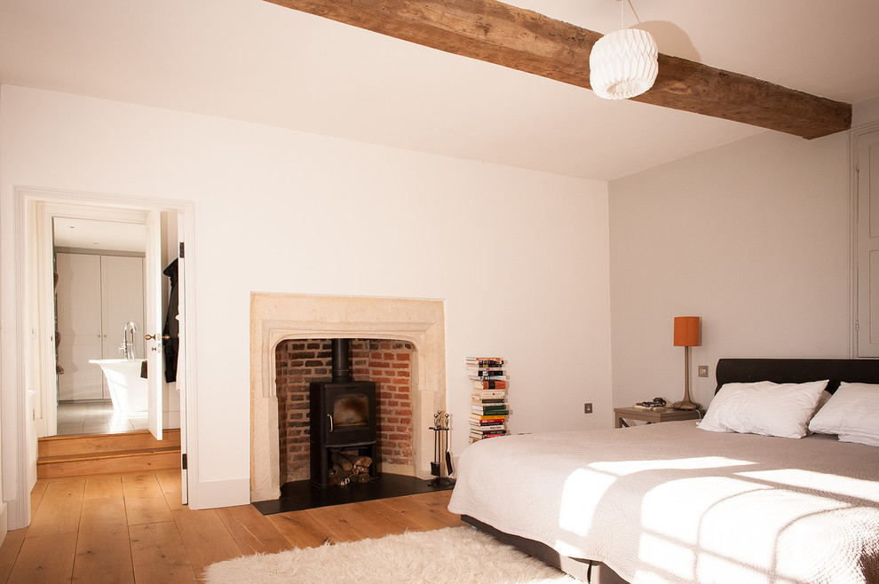 Imagen de dormitorio principal de estilo de casa de campo grande con paredes blancas, suelo de madera clara, estufa de leña y marco de chimenea de piedra