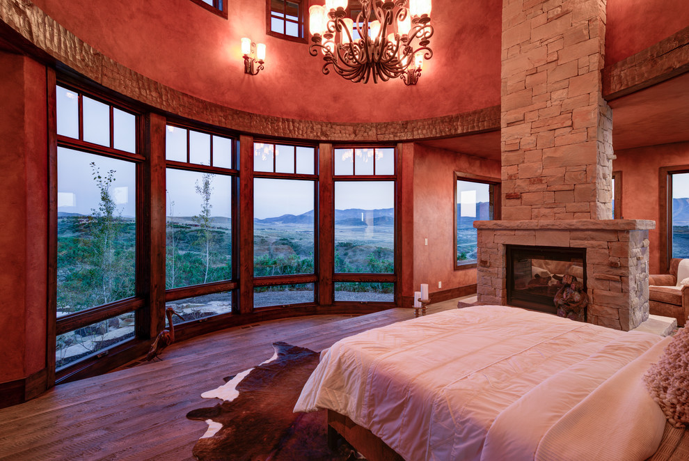 Diseño de dormitorio principal rústico grande con paredes rojas, suelo de madera en tonos medios, chimenea de doble cara, marco de chimenea de piedra y suelo marrón