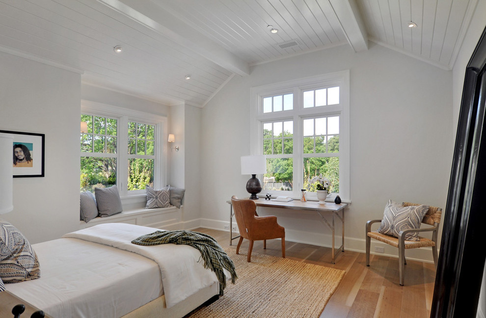 Modelo de dormitorio de estilo de casa de campo con paredes blancas, suelo de madera en tonos medios y con escritorio