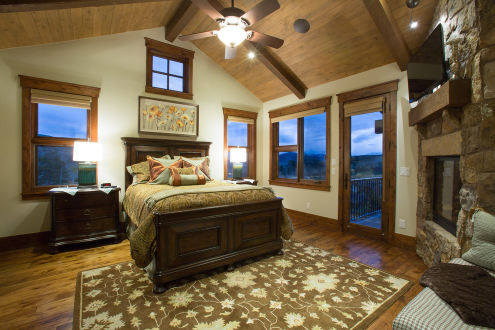 Ejemplo de dormitorio principal rural grande con paredes beige, suelo de madera oscura, chimenea de esquina, marco de chimenea de piedra y suelo marrón