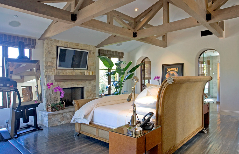 This is an example of a large mediterranean master bedroom in Los Angeles with white walls, dark hardwood flooring, a standard fireplace and a stone fireplace surround.