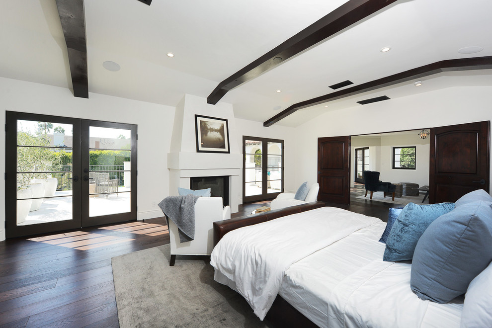 Großes Mediterranes Hauptschlafzimmer mit weißer Wandfarbe, dunklem Holzboden, Kamin, Kaminumrandung aus Beton und braunem Boden in Los Angeles