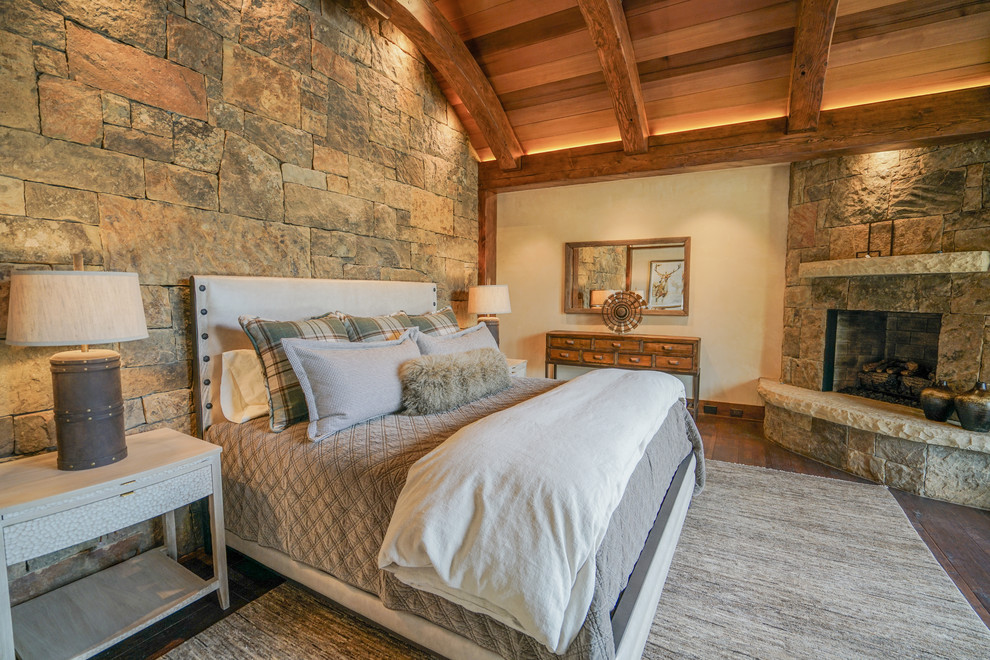 Diseño de dormitorio principal rural grande con paredes beige, suelo de madera oscura, chimenea de esquina, marco de chimenea de piedra y suelo marrón
