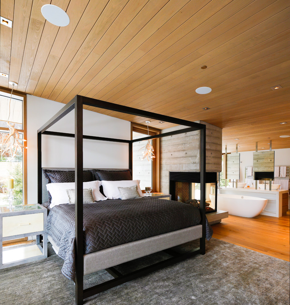 Ejemplo de dormitorio principal rústico grande con paredes marrones, suelo de madera clara, chimenea de doble cara y marco de chimenea de madera