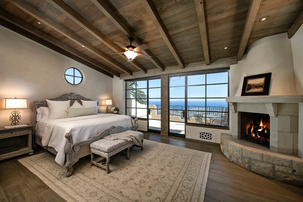 Mittelgroßes Mediterranes Hauptschlafzimmer mit beiger Wandfarbe, dunklem Holzboden, Eckkamin, Kaminumrandung aus Stein und braunem Boden in Santa Barbara
