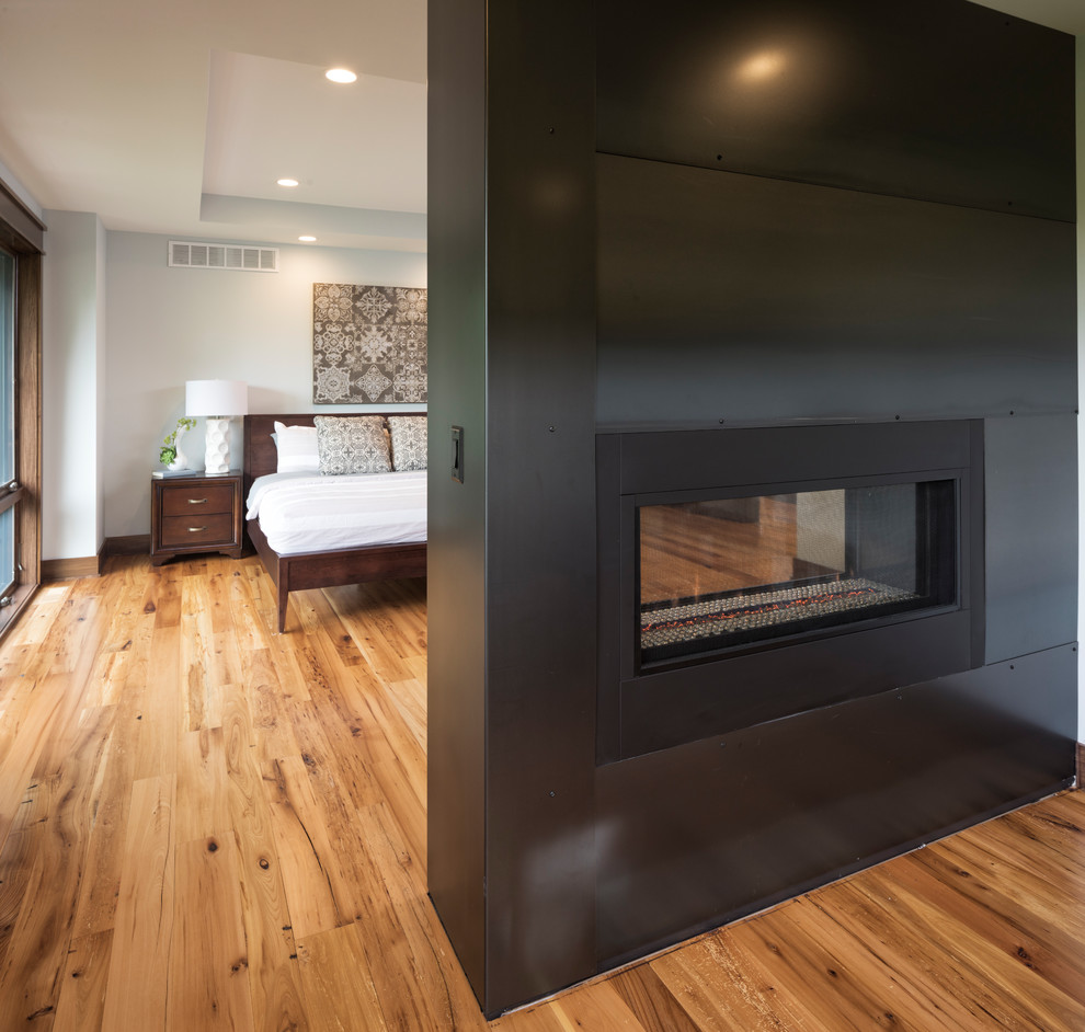 Imagen de dormitorio principal moderno con suelo de madera en tonos medios, chimenea de doble cara y marco de chimenea de metal