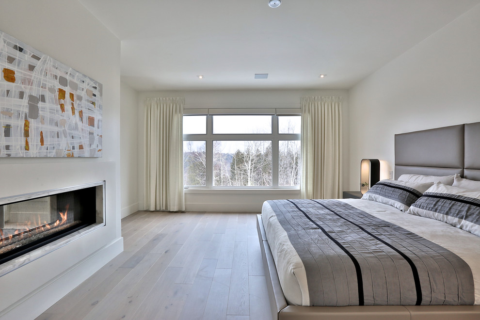 Ejemplo de dormitorio principal actual extra grande con paredes blancas, suelo de madera clara, chimenea lineal y marco de chimenea de piedra