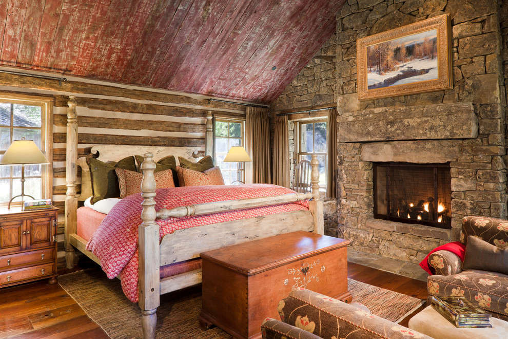 Bild på ett rustikt sovrum, med en spiselkrans i sten, grå väggar, mellanmörkt trägolv, en standard öppen spis och brunt golv
