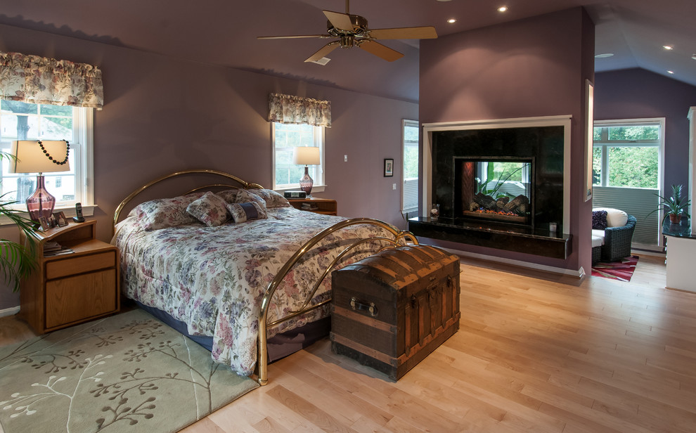 Ejemplo de dormitorio principal de estilo americano grande con paredes púrpuras, suelo de madera clara y chimenea de doble cara